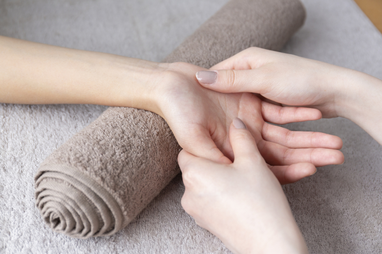 массаж кисти руки, лежащей на свернутом бежевом махровом полотенце