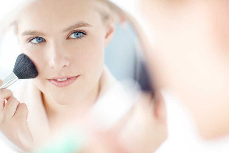 голубоглазая девушка смотрит в зеркало и наносит пудру с помощью косметической кисти