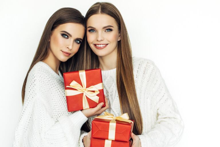 две девушки в белых длинных свитерах, с ухоженной кожей и длинными гладкими волосами держат красные коробки с подарками