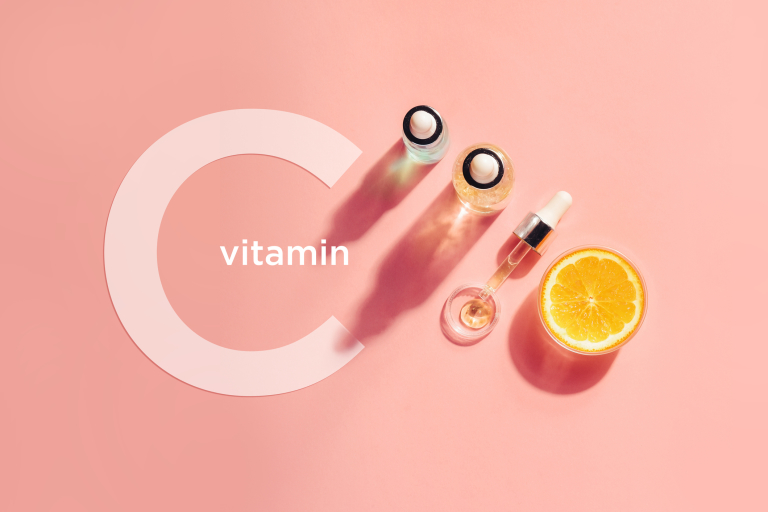 Витамин С на розовом фоне в ряд выставлены бутылочки с сыворотками, ппетка и кружок апельсина