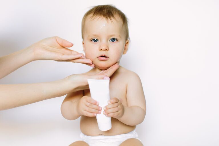 Младенец держит тюбик с кремом, а мамины руки ухаживают за кожей ребенка