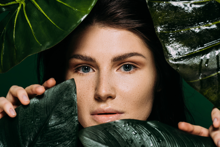 Девушка с органическим кремом на лице выглядывает из-за зеленых листьев фикуса