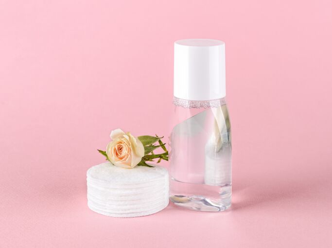 Прозрачная бутылка с тоником для жирной кожи, бутон розы и ватные диски, розовый фон