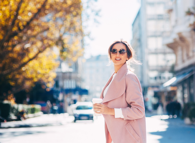 Молодая женщина в солнечных очках и розовом пальто на фоне городского пейзажа