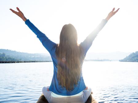 девушка в голубой кофте и белых штанах сидит спиной к зрителю и подняв руки, лицом к восходящему солнцу на озере