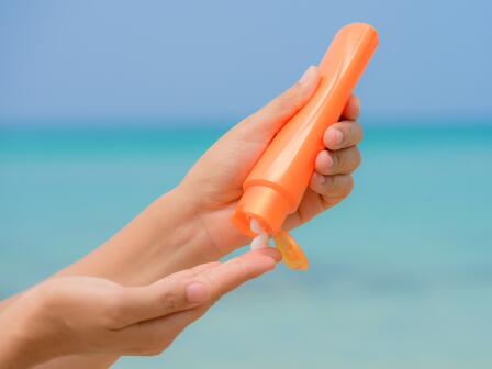 Крупный план рук, выдавливающих крем из оранжевой бутылочки на фоне моря