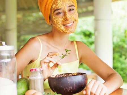 девушка в оранжевом тюрбане из полотенца и желтом топике держит на лице самодельную домашнюю маску желтого цвета, на столе перед ней продукты для домашней косметики
