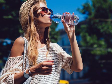 девушка в полосатом топе в соломенной шляпе и солнцезащтных очках пьет воду на улице